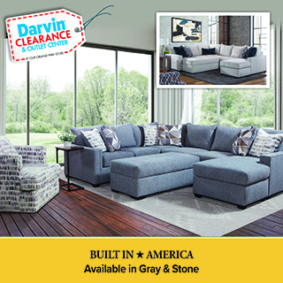 An advertisement for Darvin Clearance & Outlet Center featuring a large blue sectional sofa with an ottoman and chaise lounge. The sofa is decorated with patterned accent pillows and sits in a room with large windows overlooking a green landscape. An inset image shows the same sectional in a light gray color. A yellow banner at the bottom reads: BUILT IN AMERICA, Available in Gray & Stone.