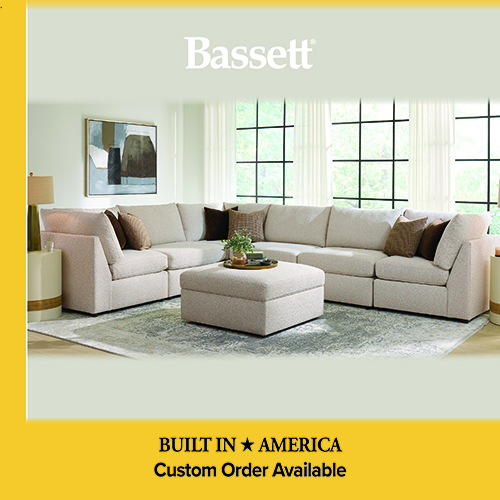 An advertisement for Bassett furniture featuring a large, L-shaped sectional sofa in a light cream fabric with several brown and patterned accent pillows. A matching square ottoman sits in the center of the sectional on a distressed gray and blue area rug. The setting is a bright room with large black-framed windows in the background. A yellow banner at the bottom reads: BUILT IN AMERICA, Custom Order Available.