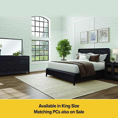 A modern bedroom furniture set featuring a black bed frame with a vertical-slatted headboard, a matching six-drawer dresser with a large mirror, and a small nightstand. The bed is dressed with white and brown bedding. The set is arranged on a light beige rug in a room with white brick walls and a large arched window. A yellow banner at the bottom reads: Available in King Size, Matching PCs also on Sale.