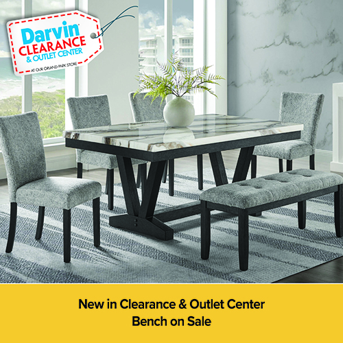 An advertisement for Darvin Clearance & Outlet Center featuring a modern dining set. The set includes a rectangular table with a marbled top and black trestle base, four gray upholstered chairs, and a matching tufted bench. The furniture is set on a striped gray rug in a room with a large window and a marble-patterned wall. A yellow banner at the bottom reads: New in Clearance & Outlet Center, Bench on Sale.