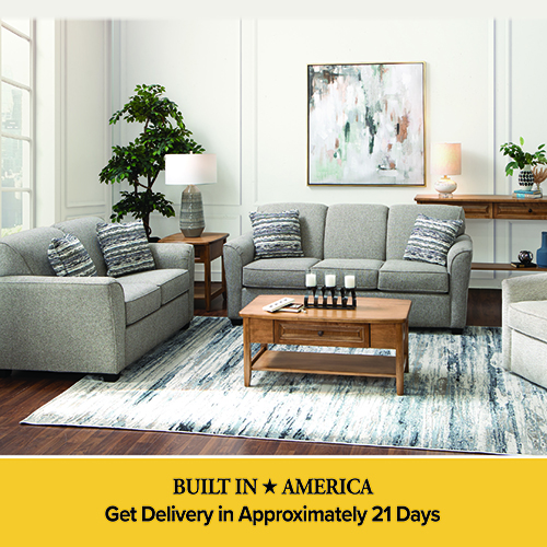 An advertisement for a living room furniture set featuring a gray sofa and matching loveseat with blue and white patterned accent pillows. The room includes a wooden coffee table and side table, set on a blue and white abstract patterned rug. A large piece of abstract art hangs on the wall in the background. A yellow banner at the bottom reads: BUILT IN AMERICA, Get Delivery in Approximately 21 Days.
