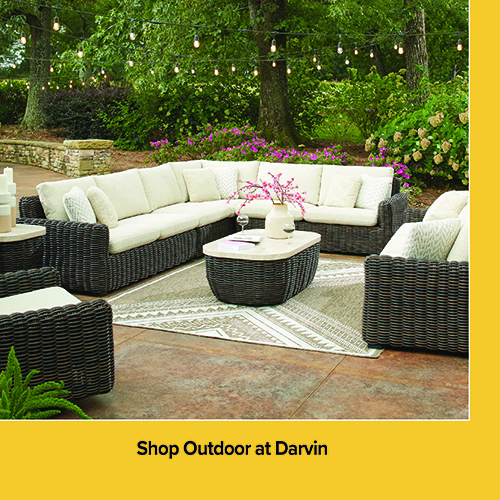 An advertisement for outdoor furniture at Darvin featuring a large, dark brown wicker sectional sofa with cream-colored cushions. The set includes a matching wicker coffee table with a light-colored top and a separate armchair, all arranged on a patterned tan and white outdoor rug. The scene is set on a stone patio under string lights with lush green trees and purple flowers in the background. A yellow banner at the bottom reads: Shop Outdoor at Darvin.