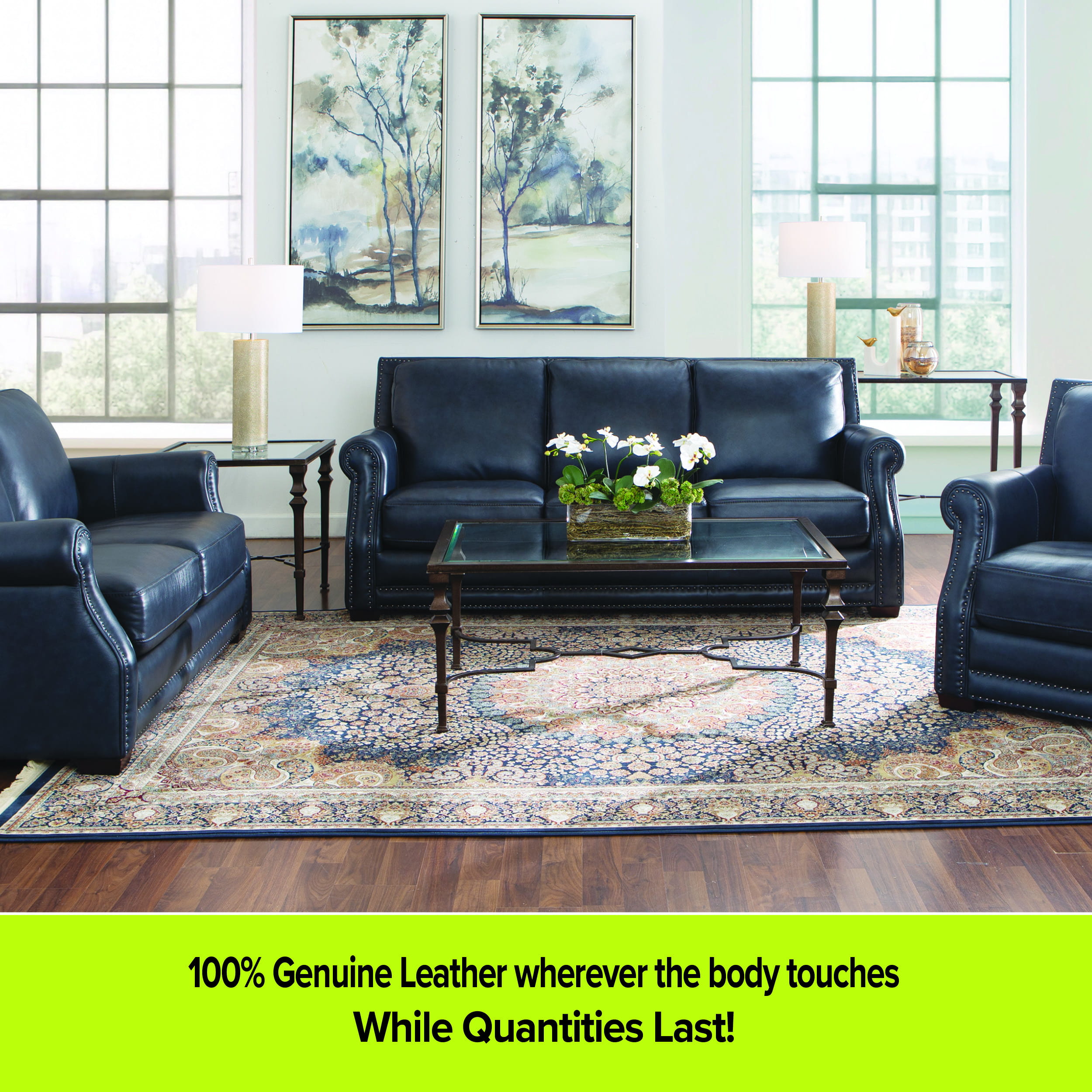 A traditional living room set featuring a navy blue leather sofa, loveseat, and armchair with rolled arms and nailhead trim. The room is styled with a large ornate oriental-style rug, a glass-top coffee table and end table, and two tall framed landscape paintings on the wall. A lime green footer reads '100% Genuine Leather wherever the body touches' and 'While Quantities Last!'.