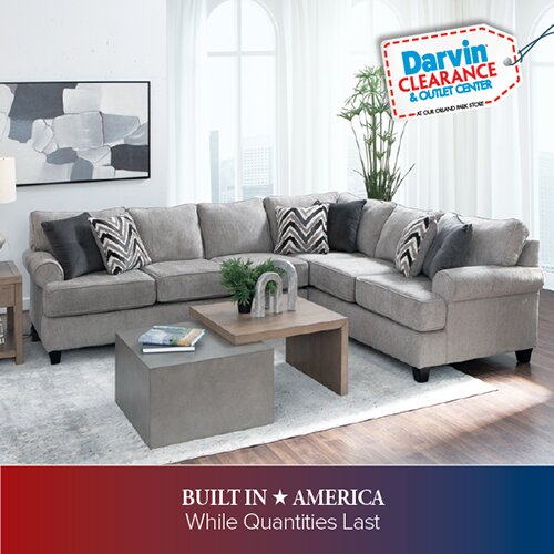 A promotional advertisement for the Darvin Clearance & Outlet Center featuring a large grey L-shaped sectional sofa with patterned accent pillows. The scene includes a modern tiered coffee table and a large indoor plant. The bottom banner reads 'BUILT IN AMERICA While Quantities Last'.