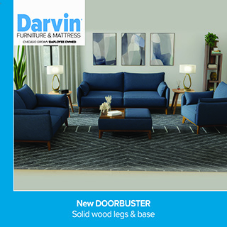 A promotional advertisement for Darvin Furniture & Mattress featuring a living room collection. The image displays a mid-century modern style living room set upholstered in vibrant blue fabric with solid wood legs and bases. The arrangement includes a large three-cushion sofa in the center, a coordinating loveseat on the left, an armchair on the right, and a matching rectangular ottoman with a wooden tray accent in the foreground. The furniture rests on a large, dark charcoal grey area rug with a white geometric chevron pattern. In the background, three abstract art pieces hang on a light grey wall, flanked by wooden end tables with unique gold loop lamps. The top left corner features the blue and black Darvin Furniture & Mattress logo with the text 'Chicago Grown Employee Owned'. A vibrant blue border frames the left side, and a matching solid blue banner along the bottom reads 'New DOORBUSTER' followed by 'Solid wood legs & base' in clean white text.