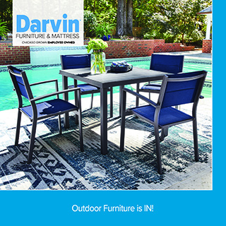 A promotional advertisement for Darvin Furniture & Mattress featuring outdoor patio furniture. The image showcases a black square outdoor dining table set with a glass vase of green hydrangeas. Surrounding the table are four matching modern outdoor dining chairs with dark metal frames and vibrant blue mesh sling seats and backs. The dining set is arranged on a blue and grey distressed geometric area rug on a concrete pool deck, right next to a bright turquoise swimming pool. In the background, lush green trees and a brick retaining wall are visible. The top left corner features the blue and black Darvin Furniture & Mattress logo with the text 'Chicago Grown Employee Owned'. A solid blue border frames the right side, and a matching solid blue banner along the bottom reads 'Outdoor Furniture is IN!' in clean white text.