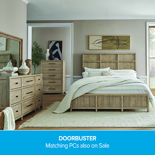 A promotional advertisement for a bedroom furniture set on sale. The image showcases a rustic or transitional style wooden bedroom collection with a light natural or weathered finish. The set includes a bed frame with a high, grid-paneled headboard and a matching low-profile footboard, dressed in white and light green striped linens. On the left is a wide nine-drawer dresser topped with decorative ceramic vases and an attached rectangular mirror. In the background, a matching tall five-drawer chest sits next to an open doorway. The furniture rests on a light-colored woven area rug with fringed edges over a hardwood floor. A solid blue banner running along the bottom reads 'DOORBUSTER' followed by 'Matching PCs also on Sale' in clean white text.