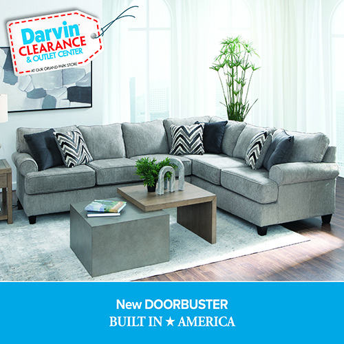 A promotional advertisement for Darvin Clearance & Outlet Center featuring a sectional sofa on sale. The image showcases a large L-shaped sectional sofa upholstered in a textured light grey fabric with rolled arms. The sofa is styled with alternating solid dark charcoal grey pillows and black-and-white chevron-patterned throw pillows. In front of the sectional rests a nesting coffee table set consisting of a square concrete-look block table and a wooden C-shaped accent table. The arrangement sits on a light grey distressed area rug over a dark hardwood floor in a bright room with tall windows and a potted green plant in the corner. In the upper left corner, a white price tag graphic with a red stitched border reads 'Darvin CLEARANCE & OUTLET CENTER AT OUR ORLAND PARK STORE'. A solid blue banner along the bottom reads 'New DOORBUSTER' followed by 'BUILT IN ★ AMERICA' in bold white text.