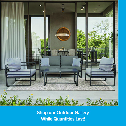 A promotional advertisement for an outdoor patio furniture collection. The image displays a modern outdoor conversation set with black metal frames and grey cushions, arranged on a light patterned outdoor area rug. The set includes a two-cushion loveseat styled with two light blue accent pillows, two matching wide armchairs, and a rectangular black metal coffee table in the center. The seating area is positioned on a concrete patio in front of a large glass-walled building that reveals an indoor dining table and a large circular gold wall accent. A solid blue border frames the right side, and a matching solid blue banner along the bottom reads 'Shop our Outdoor Gallery' followed by 'While Quantities Last!' in clean white text.