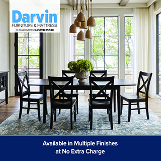 A promotional advertisement for a dining room furniture collection at Darvin Furniture & Mattress. The image features a long, dark espresso-finished rectangular wooden dining table surrounded by six matching wooden chairs with distinctive X-back designs. In the center of the table sits a large stone urn filled with vibrant green hydrangeas. The dining set is arranged on a large abstract blue, cream, and grey patterned area rug over light hardwood floors. Above the table hangs a modern multi-light pendant fixture with bell-shaped ceramic shades. The room is bright and open, characterized by tall grid-style windows with white curtains that offer a view of greenery outside. The white Darvin Furniture & Mattress logo is positioned in the upper left corner. A solid dark blue banner runs across the bottom with white text that reads, 'Available in Multiple Finishes' stacked above 'at No Extra Charge'.