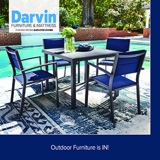 A promotional advertisement for an outdoor patio collection at Darvin Furniture & Mattress. The graphic highlights a modern outdoor dining set featuring a square black table surrounded by four matching metallic armchairs with royal blue mesh fabric seats and backs. In the center of the table sits a clear glass vase filled with green flowers. The dining set is arranged on a blue, grey, and cream patterned outdoor area rug next to a clear blue backyard swimming pool. In the background, a low brick wall separates the pool area from a lush green lawn and mature trees. The white Darvin Furniture & Mattress logo is positioned in the upper left corner. A solid dark blue banner running along the bottom of the graphic features white text that reads: 'Outdoor Furniture is IN!'.
