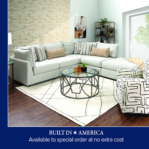 A promotional advertisement graphic for an American-built living room collection. The image showcases a large, light grey or off-white fabric sectional sofa arranged in an L-shape with an attached chaise lounge on the right end. The sectional is styled with multiple accent pillows in various patterns, including vertical stripes, brown textured fabric, and a black-and-white geometric line motif. In front of the sofa sits a round coffee table with a glass top and a dark metal frame featuring an interlocking open geometric design. The furniture rests on a cream-colored area rug with thin black geometric lines over dark hardwood floors. To the right, an armchair upholstered in the same black-and-white geometric line pattern is partially visible. The room features a light-colored brick accent wall on the left, a light blue wall in the background decorated with abstract artwork, and a tall metal shelving unit filled with decor pieces. A solid dark blue banner runs along the bottom of the graphic, featuring white text that reads 'BUILT IN ★ AMERICA' stacked above 'Available to special order at no extra cost'.