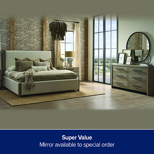 A promotional advertisement graphic for a bedroom furniture collection. The image showcases a large upholstered platform bed in a light cream or beige fabric, styled with multiple earth-toned accent pillows and a textured throw blanket. The bed rests on a tan woven area rug over light hardwood floors. To the right, a matching light wood six-drawer dresser with dark metal handles is displayed, topped with small framed pictures and a large round black-framed mirror. The room features a rustic stone accent wall with vertical wooden support beams behind the bed, a matching nightstand with a table lamp in the corner, and large, tall grid-style windows and doors looking out onto a landscape. A solid dark blue banner runs along the bottom of the graphic, featuring white text that reads 'Super Value' stacked above 'Mirror available to special order'.