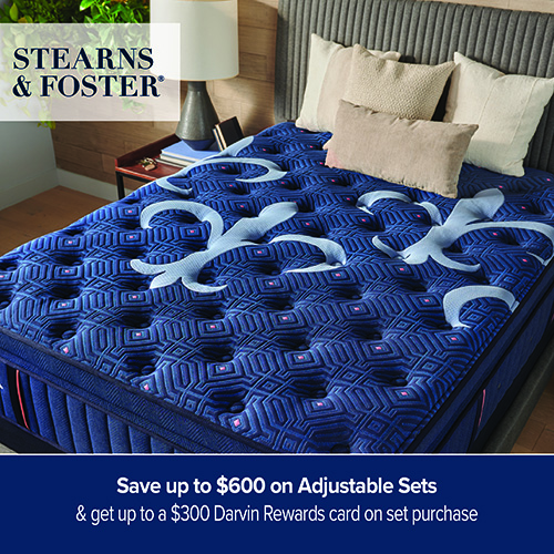 A promotional advertisement graphic for a Stearns & Foster mattress sale. The image showcases a thick luxury mattress featuring an intricate geometric quilted pattern in deep royal blue with red accents, overlaid with large, light blue fleur-de-lis tufted motifs. The mattress is displayed on a bed frame with a textured dark grey fluted headboard, styled with several neutral-toned pillows. In the background, a small wooden nightstand holds a sleek table lamp and books. The Stearns & Foster logo is positioned in a white block in the upper left corner. A solid dark blue banner runs along the bottom of the graphic, featuring white text that reads 'Save up to $600 on Adjustable Sets' stacked above '& get up to a $300 Darvin Rewards card on set purchase'.