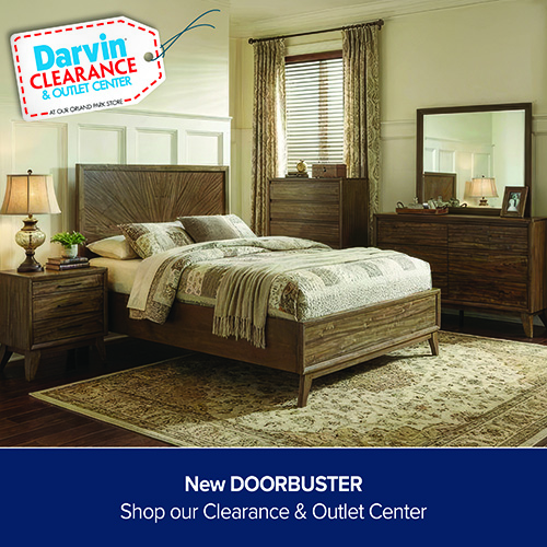 A promotional advertisement graphic for a bedroom furniture collection featured at the Darvin Clearance & Outlet Center. The image showcases a complete rustic medium-brown wooden bedroom suite, including a queen or king-sized bed frame with a high headboard featuring a decorative starburst or sunburst wood grain pattern. The bed is styled with a neutral patchwork-patterned quilt and a ribbed green throw blanket, resting on a large patterned traditional area rug over dark hardwood floors. The set includes a matching three-drawer nightstand with a classic table lamp on the left, a tall chest of drawers in front of a window with blinds and patterned curtains in the background, and a wide six-drawer dresser topped with a large square matching wooden-framed mirror on the right. The walls feature light cream paint with white wainscoting panels. In the upper left corner, a white clearance price tag graphic with a red dashed border reads 'Darvin CLEARANCE & OUTLET CENTER'. A solid dark blue banner runs across the bottom with white text that reads, 'New DOORBUSTER' stacked above 'Shop our Clearance & Outlet Center'.