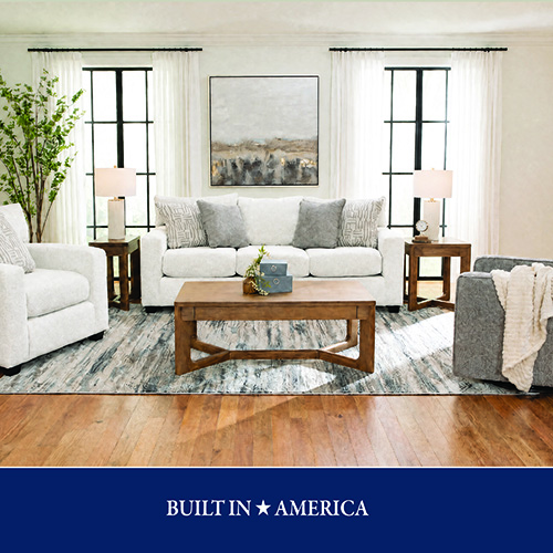 A promotional advertisement graphic for an American-built living room collection. The image features a bright, symmetrical living room arrangement showcasing a textured off-white three-cushion sofa styled with grey and patterned accent pillows. In front of the sofa sits a rectangular medium-toned wooden coffee table with a geometric base design. The setup includes a matching off-white armchair on the left and a textured grey armchair with a plush cream throw blanket draped over it on the right. Two matching wooden end tables with cross-brace bases and white cylinder table lamps flank the sofa. The entire seating group is arranged on a large abstract blue, grey, and cream patterned area rug over warm hardwood floors. In the background, two large grid-style windows with sheer white curtains flank a large square piece of abstract artwork in neutral earth tones. A solid dark blue banner runs along the bottom of the graphic, featuring white text that reads 'BUILT IN ★ AMERICA'.