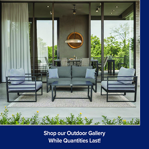 A promotional advertisement graphic for an outdoor living collection. The image showcases a modern patio conversation set with slim, black metal frames and slate grey cushions, including a two-cushion loveseat flanked by two matching deep-seating armchairs. A matching black rectangular coffee table sits in the center of the arrangement, which rests on a beige and grey patterned outdoor rug. The setup is positioned on a covered concrete patio bordered by low green landscaping with small yellow flowers in the foreground. Large, open floor-to-ceiling glass sliding doors reveal an indoor dining area behind the patio set, complete with a long dining table, chairs, and a large round bronze-toned pendant light. Trees and greenery are visible through the windows in the background. A solid dark blue banner runs along the bottom of the graphic, featuring white text that reads 'Shop our Outdoor Gallery' stacked above 'While Quantities Last!'.