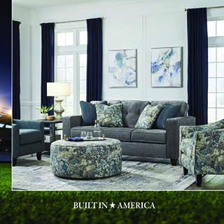 Living room with a dark gray tufted sofa, matching armchair, round patterned ottoman, wood side table with lamp, and framed art above navy blue curtains, with a stadium image accent on the left edge. Text reads BUILT IN AMERICA.