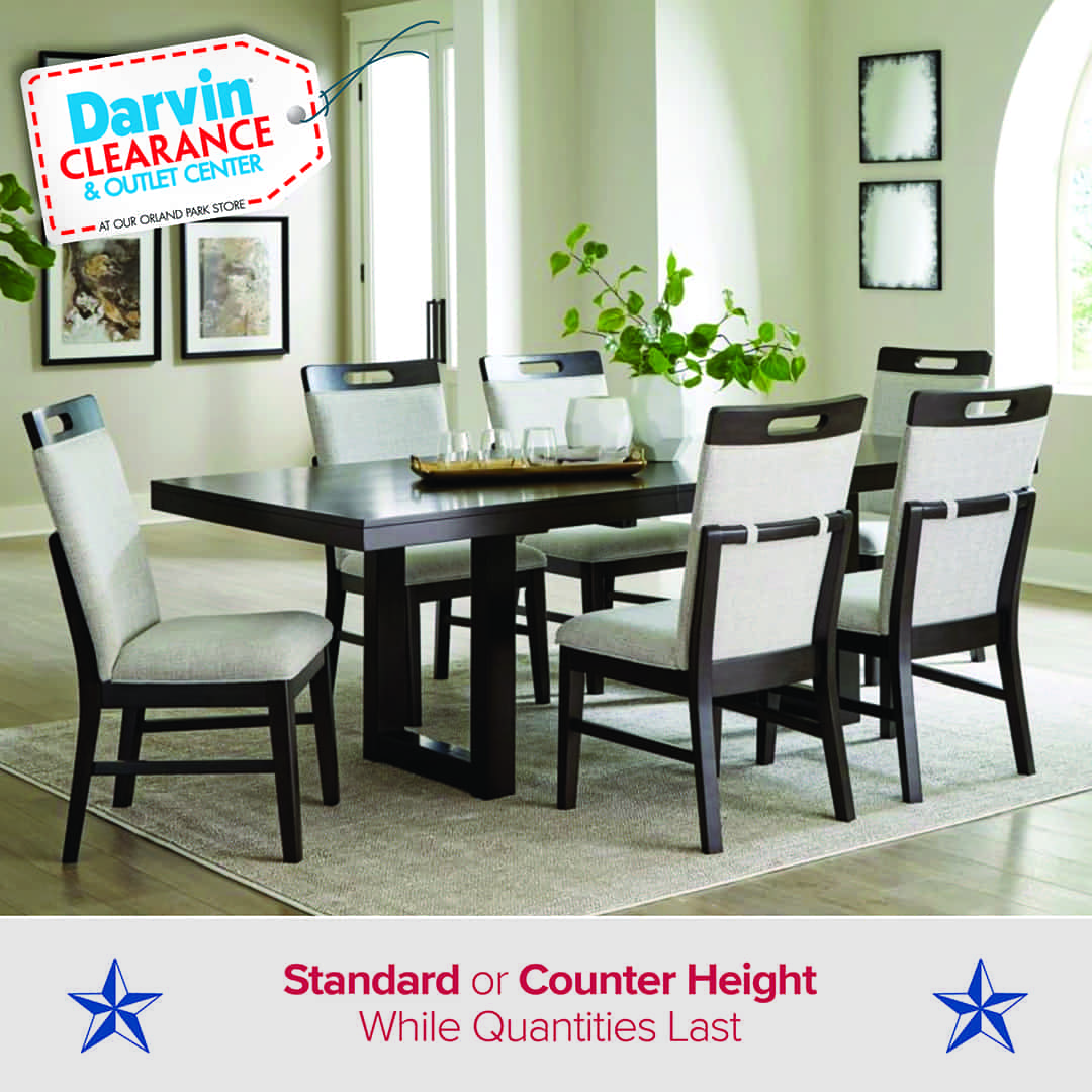 Modern dining room with a dark wood table and six upholstered chairs featuring light gray seats and backs. The space has large windows, light walls, and decorative wall art. Text on the image reads: “Darvin Clearance & Outlet Center – Standard or Counter Height – While Quantities Last,” with blue stars on each side.