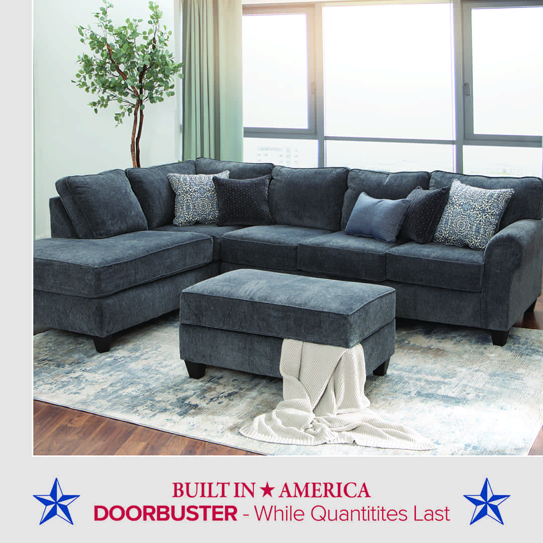 Dark gray sectional sofa with matching ottoman placed in a bright living room with large windows and light curtains. The sofa has several patterned and solid throw pillows, and a light knit blanket draped over the ottoman. A potted tree stands in the corner, and the setup rests on a light patterned area rug over hardwood floors. Text below reads: “BUILT IN AMERICA – DOORBUSTER – While Quantities Last,” with blue stars on each side.
