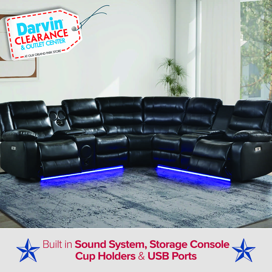 Black leather reclining sectional sofa with built-in cup holders, storage consoles, and blue LED lighting along the base. The sectional is arranged in an L-shape and placed in a bright living room with a large window and potted plants. Text on the image reads: “Darvin Clearance & Outlet Center – Built-in Sound System, Storage Console, Cup Holders & USB Ports,” with blue stars on each side.