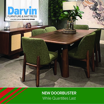 A Darvin Furniture & Mattress promotional image featuring a modern dining room setup with a wooden table and four green upholstered chairs. A sideboard with light-colored paneling sits in the background, along with a tall plant and a table lamp. The lower portion of the image includes a green banner with the text NEW DOORBUSTER — While Quantities Last.