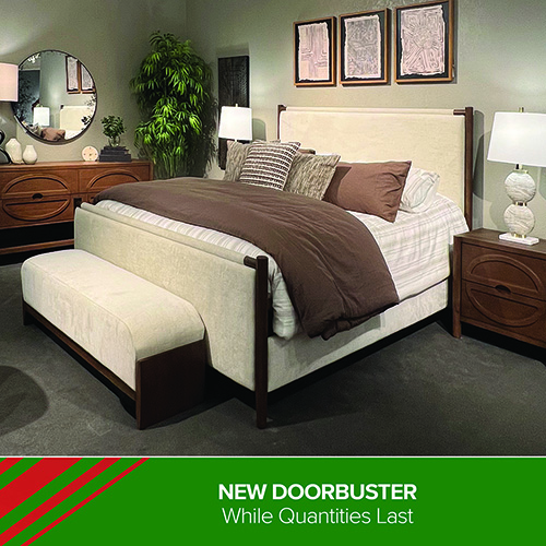 A Darvin Furniture & Mattress bedroom display featuring a modern cream-upholstered bed with wood accents and layered neutral bedding. A matching upholstered bench sits at the foot of the bed. The room includes two wooden nightstands with white lamps, a wooden dresser with a round mirror, decorative wall art, and a large potted plant. A green banner at the bottom reads: NEW DOORBUSTER — While Quantities Last.