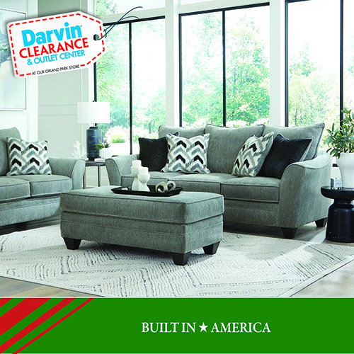 A Darvin Clearance & Outlet Center promotional image featuring a coordinated living room set in soft gray upholstery, including a sofa, loveseat, and a matching ottoman. The furniture is styled with patterned black-and-white accent pillows. The room has large floor-to-ceiling windows, a light area rug, a white table lamp, and decorative accessories on a side table. A green banner at the bottom reads: BUILT IN AMERICA.