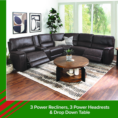 A living room scene featuring a large dark brown leather sectional sofa with multiple power recliners and power headrests. One recliner is extended, showing its functionality. The space includes a round wooden coffee table with shelves, a patterned area rug, and framed abstract art on the wall. A tall plant sits in the corner near large windows overlooking greenery. Text at the bottom reads: 3 Power Recliners, 3 Power Headrests & Drop Down Table.