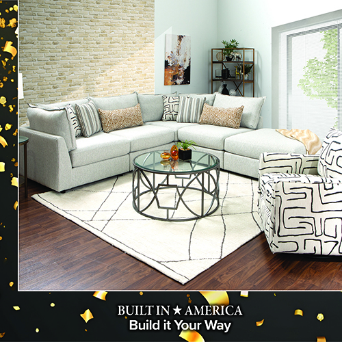 Promotional image of a contemporary living room featuring a dark brown leather sofa and matching seating with patterned throw pillows, arranged around a wooden coffee table on a light area rug. Text at the bottom reads, Built in America, with a star icon, and Matching PCs also on Sale.