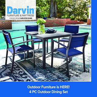 Outdoor patio furniture set featuring a dark dining table with matching cushioned chairs on a patterned rug, arranged on a covered porch with greenery in the background; inset shows a close-up of the tabletop with an umbrella hole, Darvin Furniture & Mattress logo in the top left, and text reading “Outdoor Furniture is IN!” at the bottom.