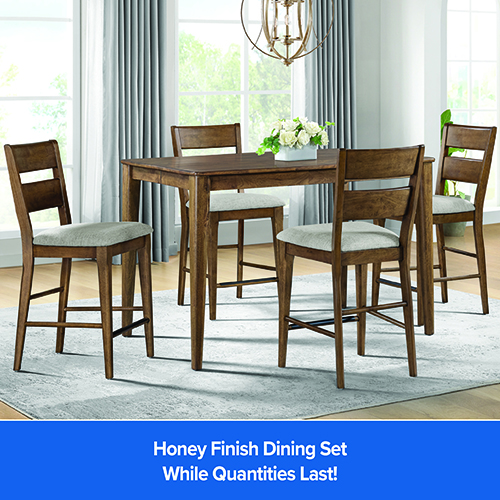 A high-angle, medium shot shows a honey-finished wood dining set for four on a light gray area rug in a bright room with large windows and light gray curtains. The rectangular table has tapered legs and a smooth top. Four matching chairs with ladder-back rests and light gray upholstered seats are arranged around the table. A white vase of white flowers sits in the center of the table. A gold, orb-shaped chandelier hangs above the table. A large window with a white frame is in the background, and a potted plant is in the corner. The bottom of the image has a blue banner with white text that reads,