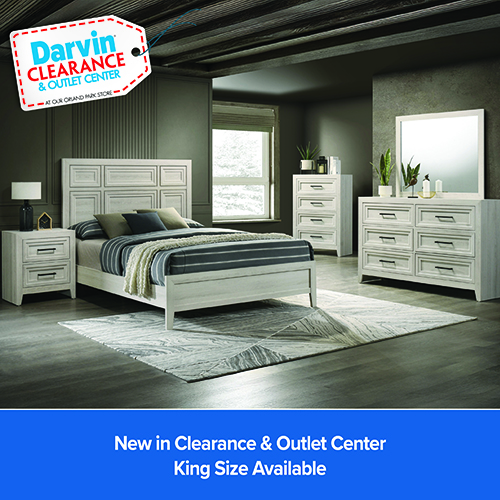 A wide shot of a contemporary bedroom set in a light-wash wood finish, situated in a room with dark olive walls and a dark wood-beamed ceiling. The set includes a bed with a paneled headboard, a two-drawer nightstand with a lamp, a five-drawer chest, and a six-drawer dresser with a large square mirror.The bed is styled with a striped navy and white comforter, neutral pillows, and a cream throw blanket. A geometric-patterned rug sits on the dark wood floor. In the top left corner, a graphic tag reads