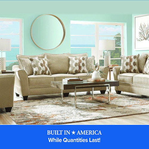 A medium shot shows a coastal-themed living room with seafoam green walls and a large window offering a view of the ocean. The room is furnished with a tan upholstered sofa and loveseat, both accented with patterned throw pillows in shades of brown, gray, and cream. A rectangular dark wood coffee table with a polished chrome base sits on a large, distressed-style area rug with gray and orange tones. Above the sofa, a large circular mirror with a gold frame is mounted on the wall. Two matching table lamps with white drum shades and textured glass bases are visible on end tables. At the bottom, a blue banner features white text that reads: