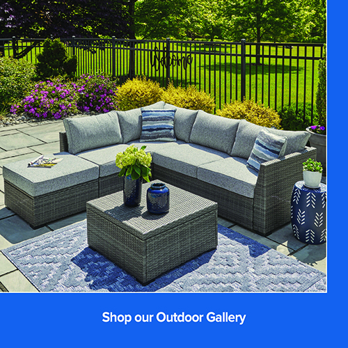 A medium, high-angle shot showcases a gray wicker sectional sofa with light gray cushions on a stone patio. The sectional is accented with blue and white striped throw pillows and includes a matching wicker ottoman. A square wicker coffee table sits on a blue patterned area rug in front of the sofa, decorated with two blue vases and green hydrangeas.In the background, a black metal fence separates the patio from a lush green lawn and trees, with a