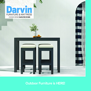 A promotional advertisement for Darvin Furniture & Mattress featuring outdoor patio furniture. The image showcases a black or dark grey counter-height outdoor console table styled with a small potted plant and a bowl of oranges. Tucked underneath the table are two matching square outdoor barstools topped with off-white cushions. The set is positioned against a minimalist white outdoor wall, with an arched doorway showing a blue and white striped curtain to the right. The upper left corner displays the blue and black Darvin Furniture & Mattress logo with the text 'Chicago Grown Employee Owned'. A solid teal banner running along the bottom reads 'Outdoor Furniture is HERE!' in clean white text.