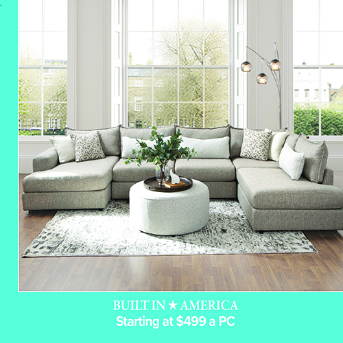 A promotional advertisement featuring a large sectional sofa. The image showcases a modern, multi-piece sectional sofa in a light grey textured fabric with dual chaise ends. It is styled with multiple coordinating throw pillows, including white solid pillows and grey and white geometric patterned pillows. A large, round upholstered ottoman sits in the center on a grey and white abstract patterned area rug. The room features a dark hardwood floor and large white-framed windows in the background. A solid teal banner along the bottom reads 'BUILT IN AMERICA' followed by 'Starting at $499 a PC' in bold white text.