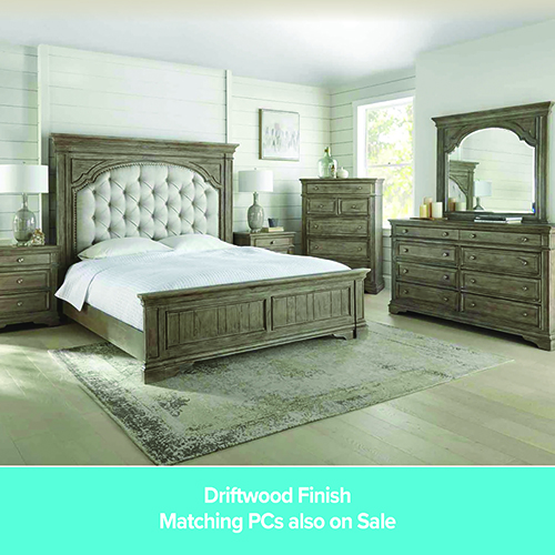 A promotional advertisement for a bedroom set in a driftwood finish. The image features a large wooden bed with a high, arched headboard featuring tufted light grey fabric upholstery. The bed is dressed in white linens and sits on a grey patterned area rug over light wood floors. Matching driftwood-finish furniture includes a nightstand with a white lamp, a tall chest of drawers, and a large dresser with an attached arched mirror. The room has light shiplap walls. A solid teal banner along the bottom reads 'Driftwood Finish' followed by 'Matching PCs also on Sale' in white text.