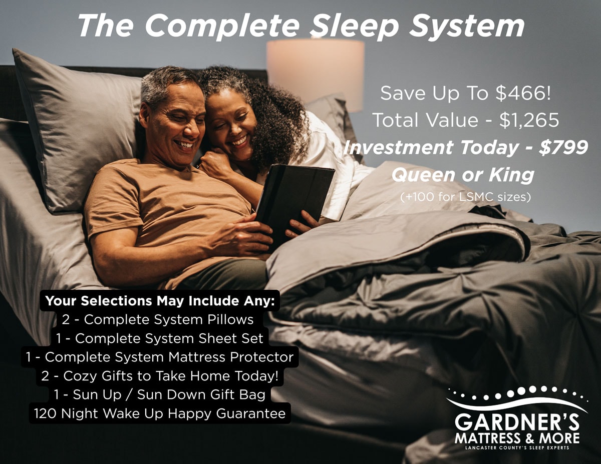 Gardner’s Mattress & More promotional graphic for The Complete Sleep System. Two people sit in bed looking at a tablet with gray bedding and pillows. Text reads: The Complete Sleep System. Save Up To $466. Total Value $1,265. Investment Today $799 Queen or King, plus $100 for LSMC sizes. Your selections may include: 2 Complete System Pillows, 1 Complete System Sheet Set, 1 Complete System Mattress Protector, 2 Cozy Gifts to Take Home Today, 1 Sun Up/Sun Down Gift Bag, and a 120 Night Wake Up Happy Guarantee. Gardner’s Mattress & More logo appears in the lower right corner.