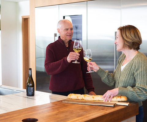 Labor Day Sale image older man and woman smiling and toasting with white wine glasses in a modern kitchen.